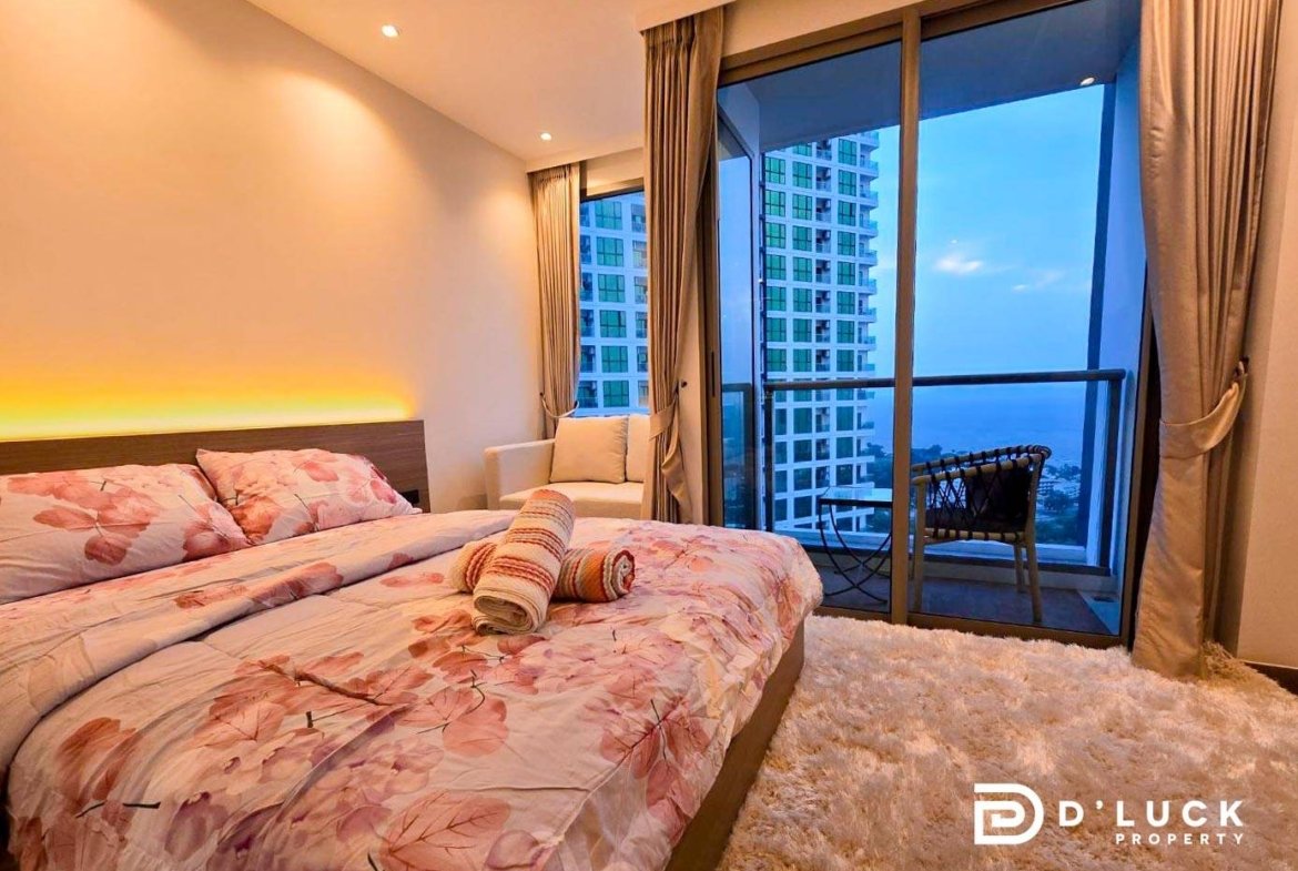 Bedroom with a double bed in pink floral bedding, rolled towels on the covers, and a balcony door opening to a city and sea view.