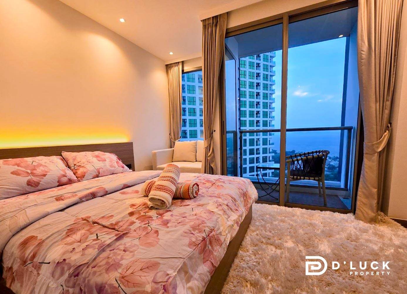 Riviera-Ocean-Drive-Studio-Rent-2609 1 Bedroom with a double bed in pink floral bedding, rolled towels on the covers, and a balcony door opening to a city and sea view.