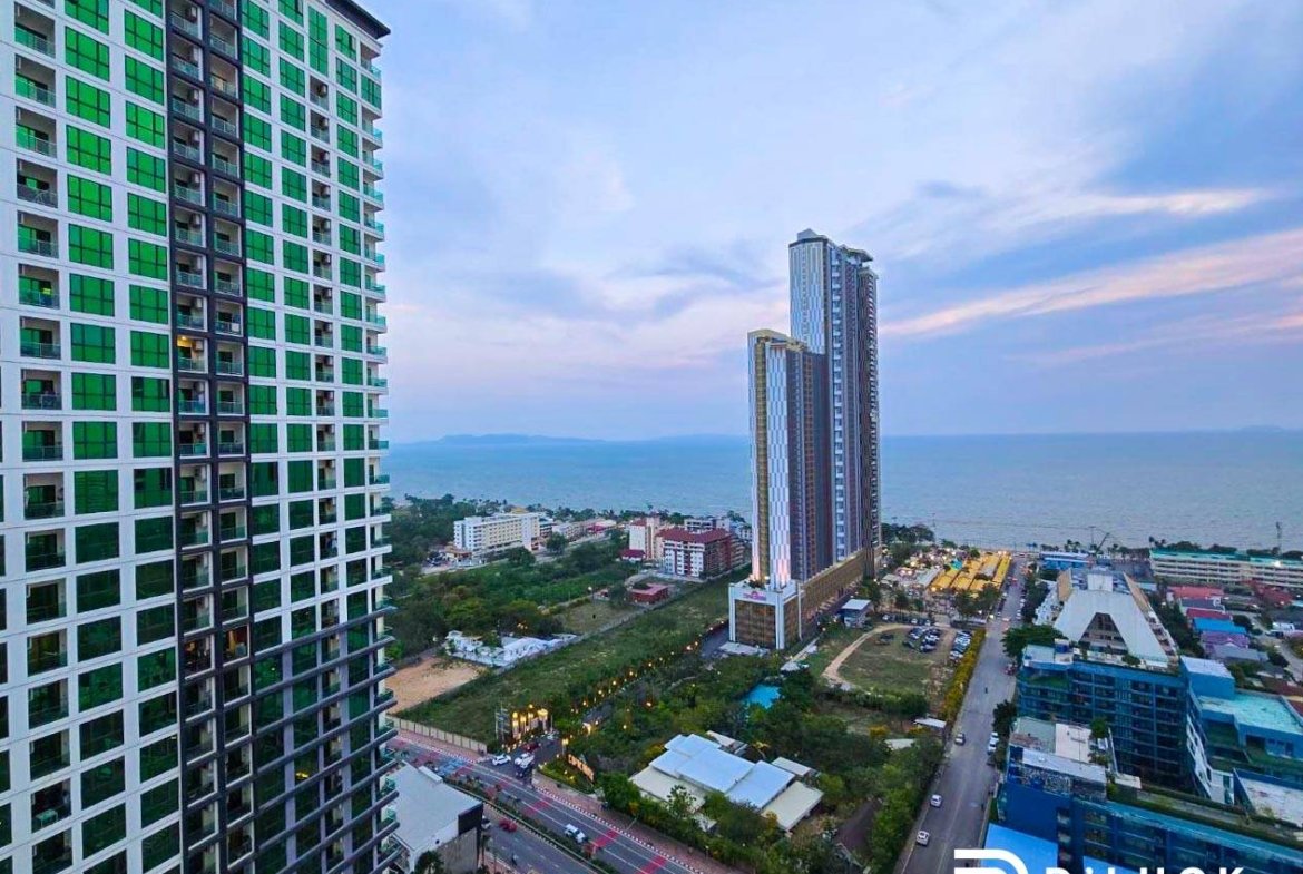 Coastal city skyline with a green-glass residential tower on the left and two tall towers near the sea, with roads and smaller buildings in the foreground.