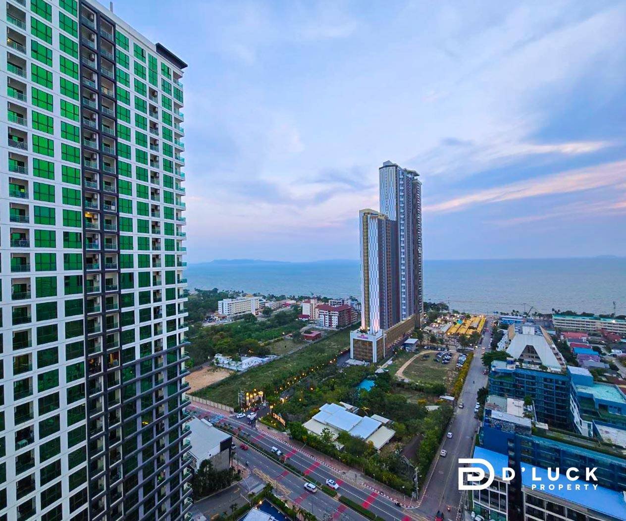 Riviera-Ocean-Drive-Studio-Rent-2609 11 Coastal city skyline with a green-glass residential tower on the left and two tall towers near the sea, with roads and smaller buildings in the foreground.