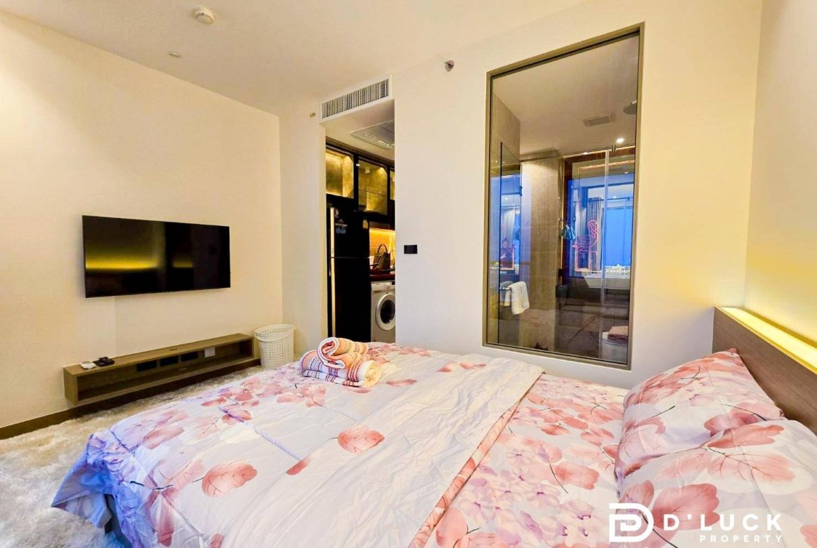 Bedroom with a pink-flowered duvet on a large bed, wall-mounted TV, and a low media console; a glass-walled bathroom and kitchenette are visible nearby.