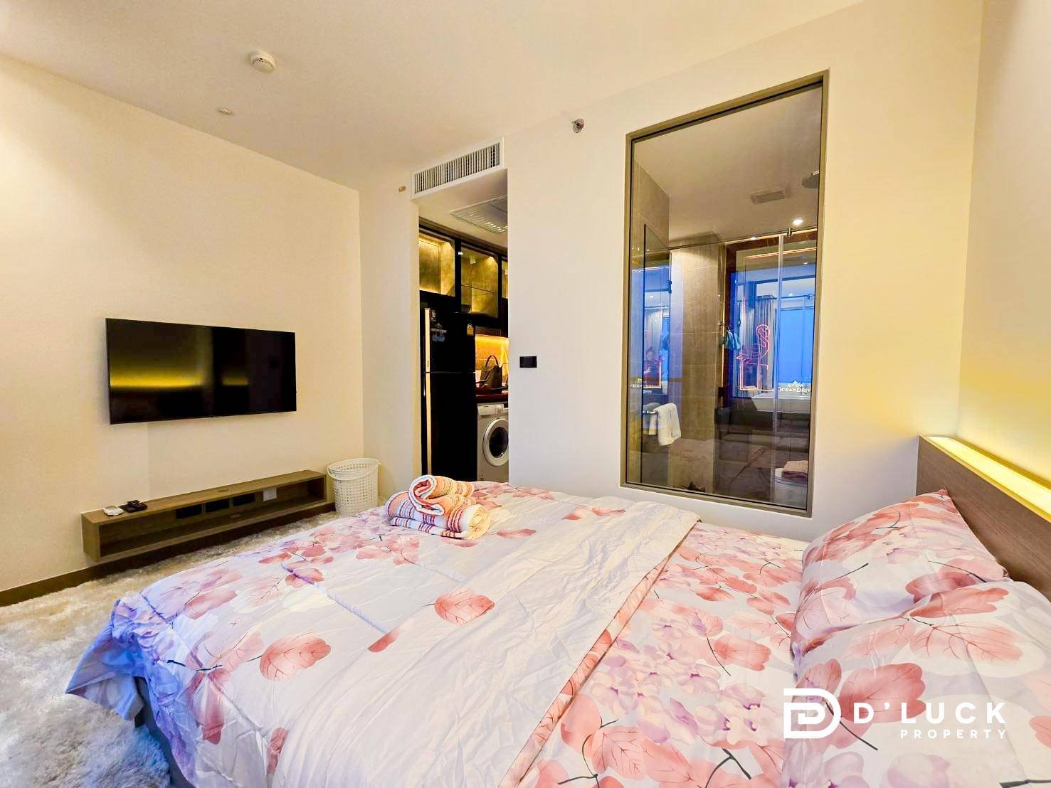 Riviera-Ocean-Drive-Studio-Rent-2609 12 Bedroom with a pink-flowered duvet on a large bed, wall-mounted TV, and a low media console; a glass-walled bathroom and kitchenette are visible nearby.