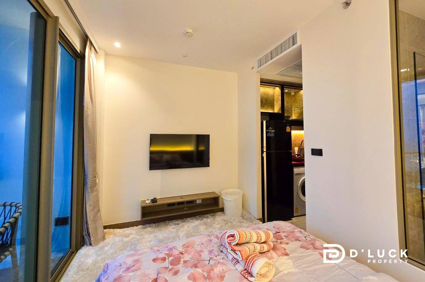 Riviera-Ocean-Drive-Studio-Rent-2609 13 Modern hotel-style bedroom with a wall-mounted TV, low media console, and a bed with pink floral bedding; rolled towels sit on the bed, and a balcony door is on the left.