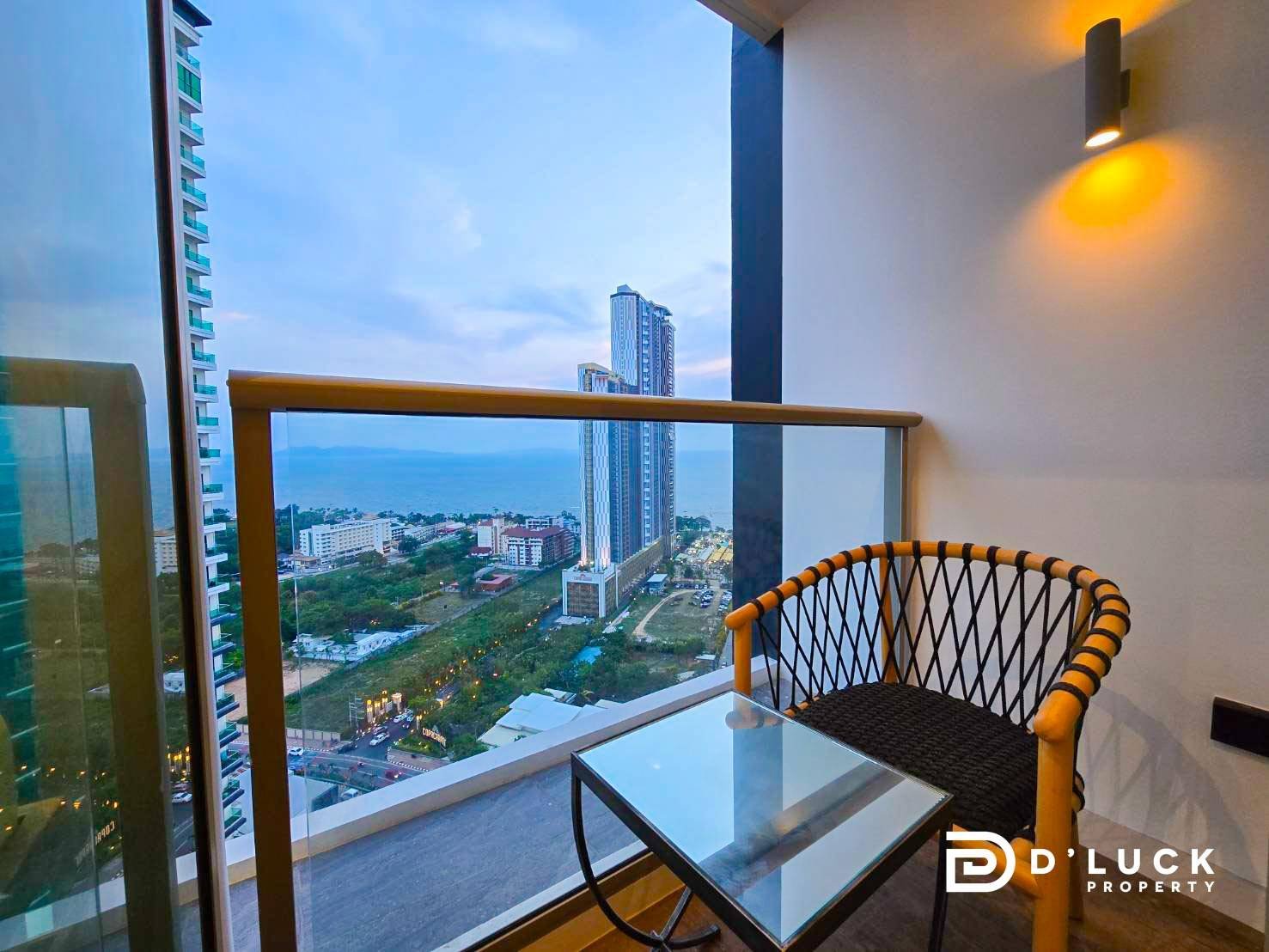 Riviera-Ocean-Drive-Studio-Rent-2609 2 Balcony seating area with a glass table and woven chair, overlooking a city and the sea horizon.