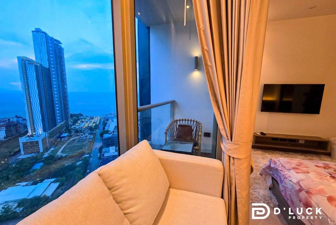 Living area in a high-rise apartment with a beige sofa facing glass balcony doors and a blue evening cityscape outside the window.