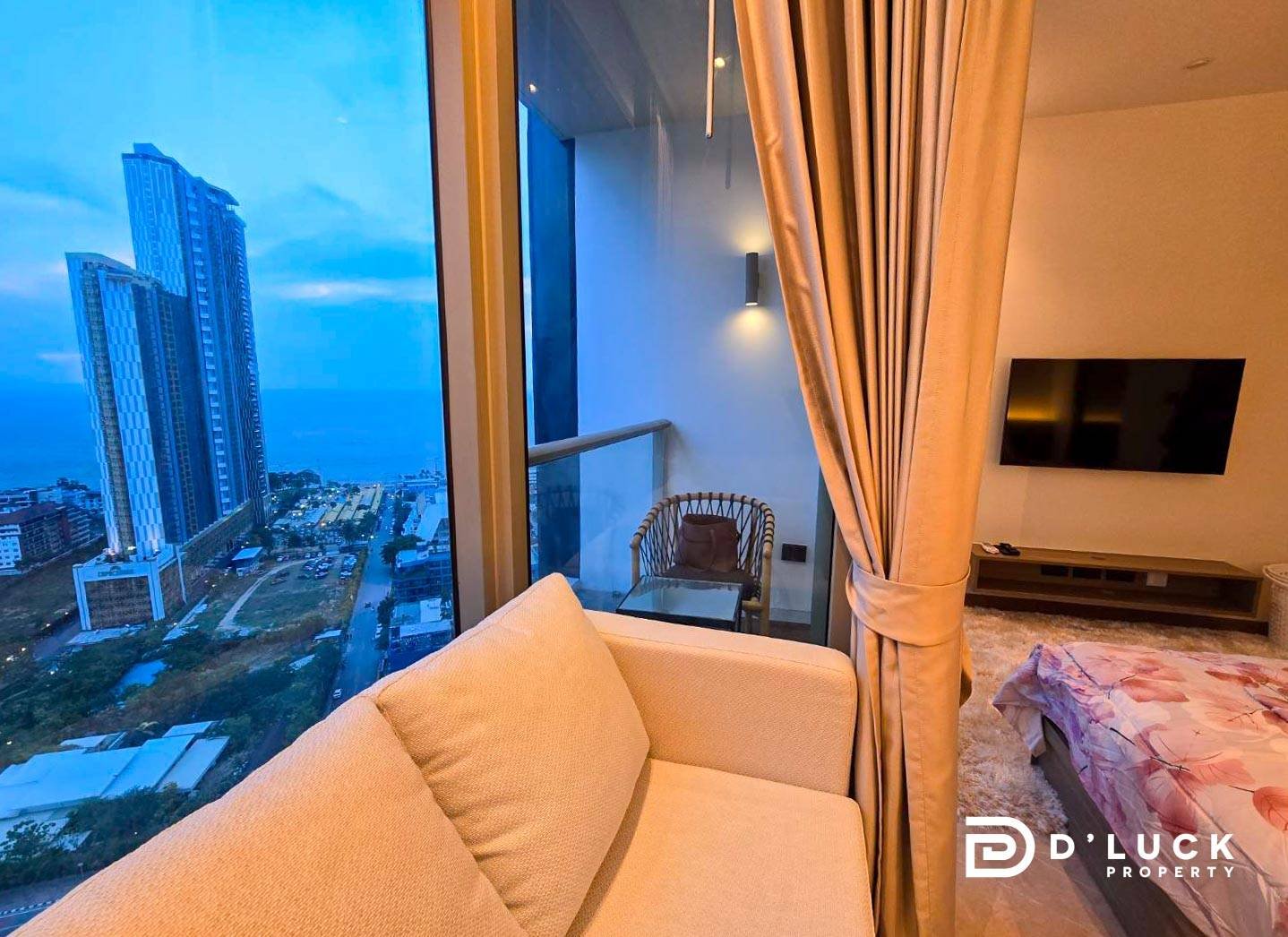Riviera-Ocean-Drive-Studio-Rent-2609 3 Living area in a high-rise apartment with a beige sofa facing glass balcony doors and a blue evening cityscape outside the window.