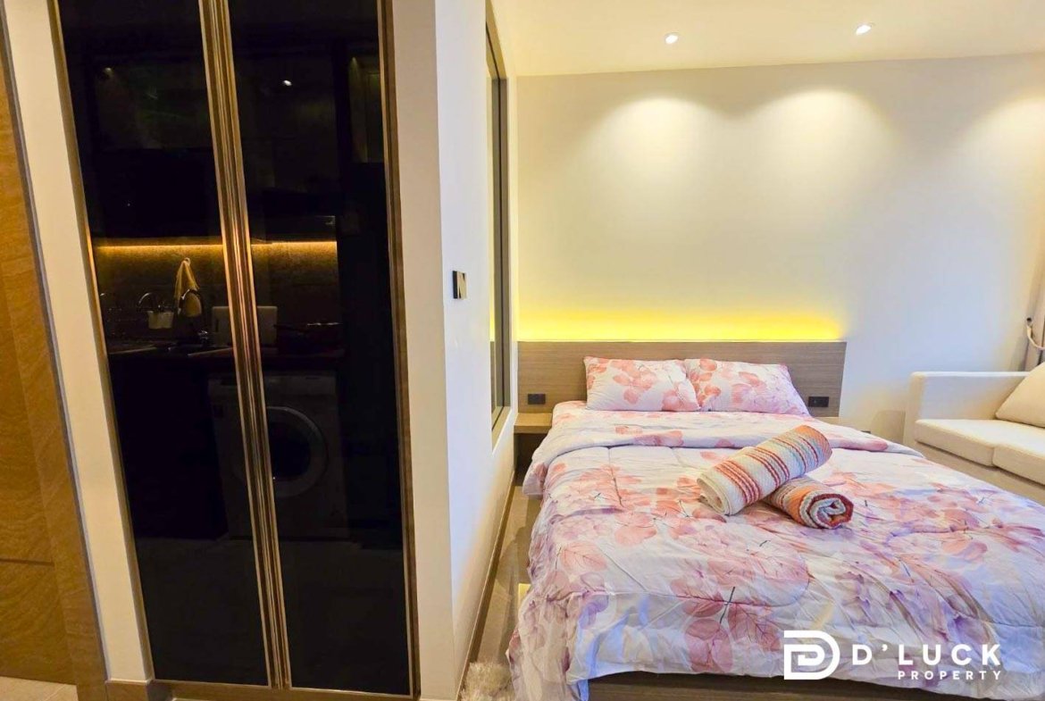 Bedroom with floral pink bedding, rolled towels on the bed, and a beige sofa alongside a mirrored wardrobe with warm lighting behind.