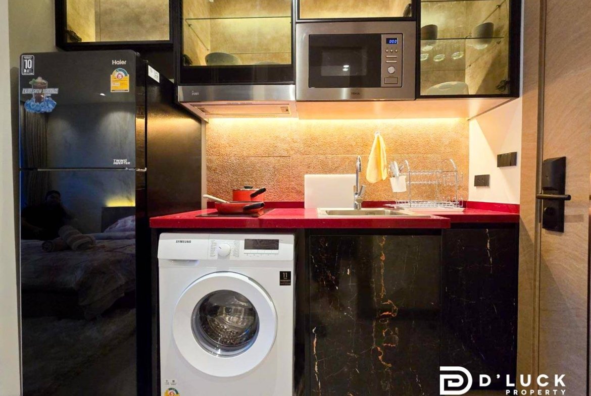 Compact kitchen with red countertop, black marble cabinets, and stainless appliances (fridge, washing machine, microwave) arranged along a single wall.