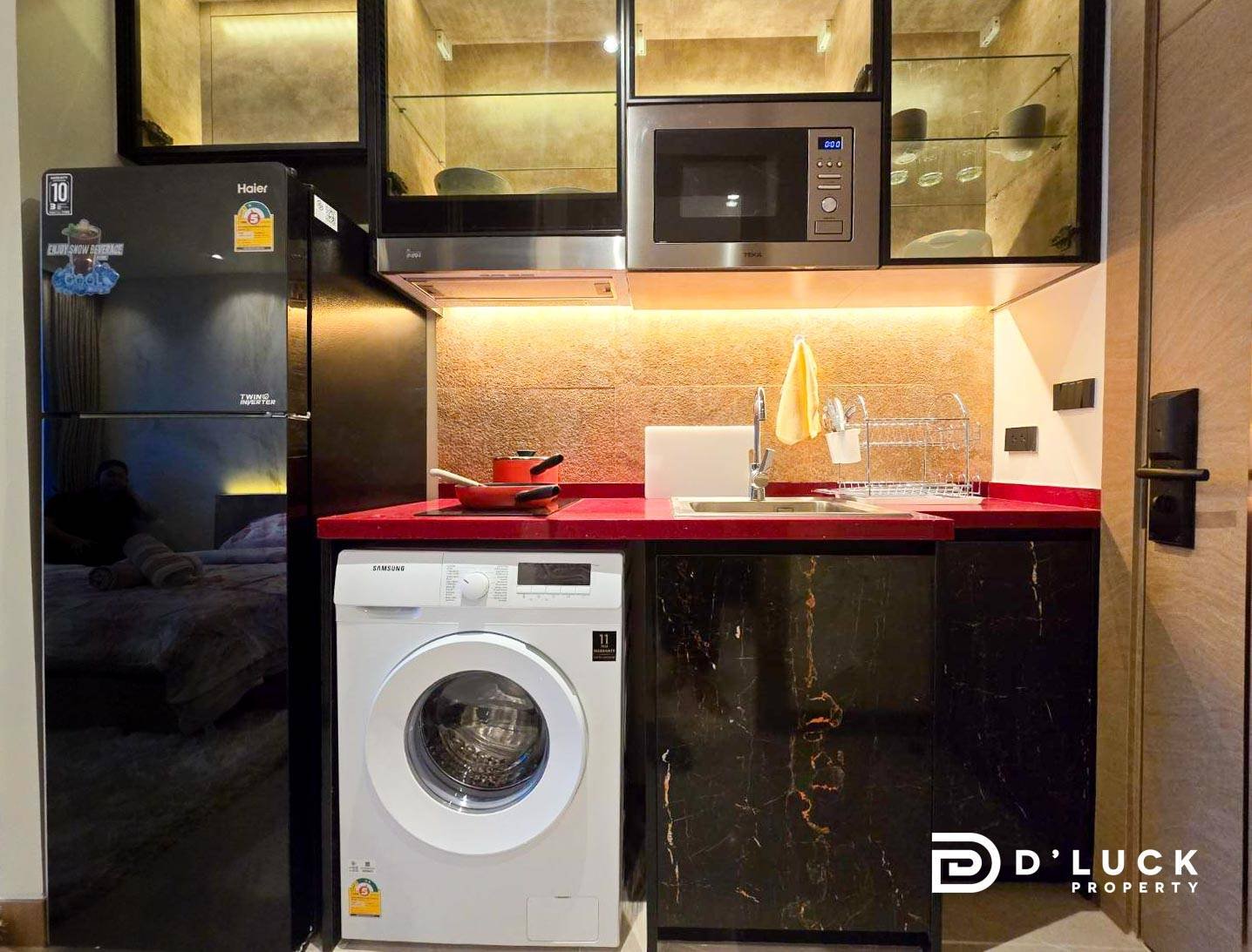 Riviera-Ocean-Drive-Studio-Rent-2609 7 Compact kitchen with red countertop, black marble cabinets, and stainless appliances (fridge, washing machine, microwave) arranged along a single wall.