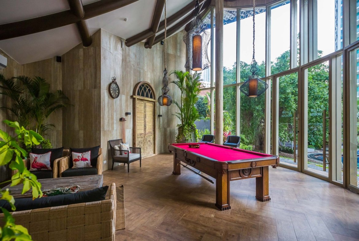 Indoor lounge with a red-felt billiards table, wicker sofas with flamingo pillows, tall glass walls, and hanging lanterns amid tropical plants and wooden ceiling beams.