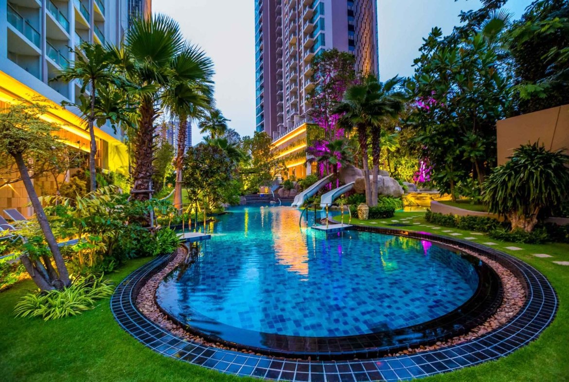 Night view of a resort pool area with palm trees, illuminated landscaping, and high-rise buildings in the background.