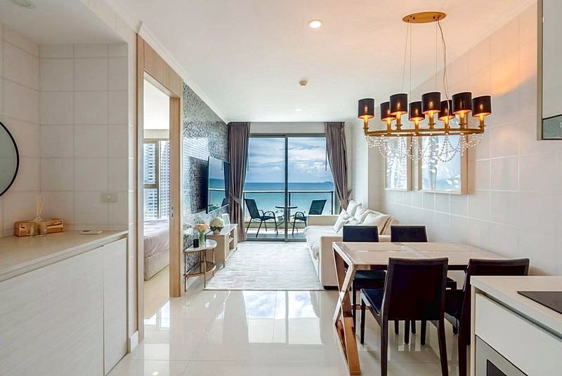 Open-plan living-dining area with a chandelier, white furniture, and a balcony overlooking the sea; bright, modern decor with a sofa and dining table by floor-to-ceiling windows.