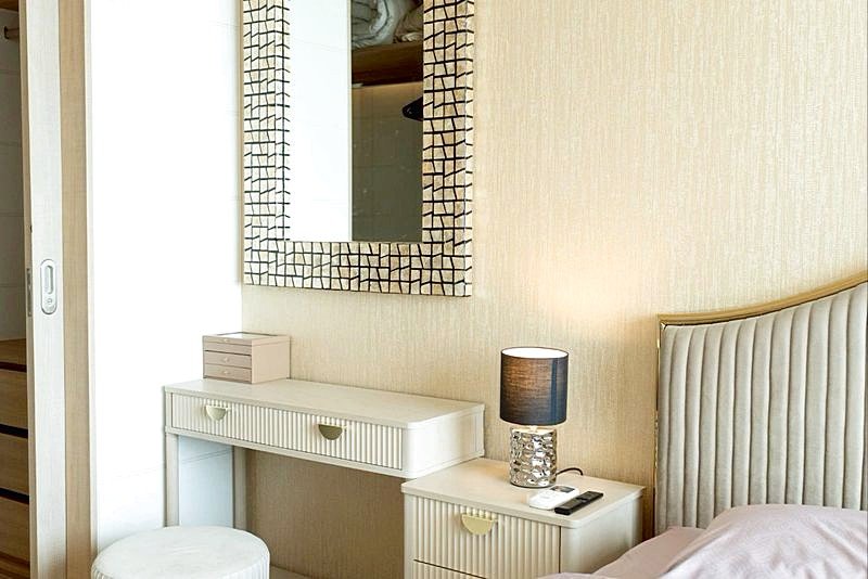 White vanity with two drawers and a mosaic-framed mirror, a small box on top, and a lamp on the nightstand beside the bed.