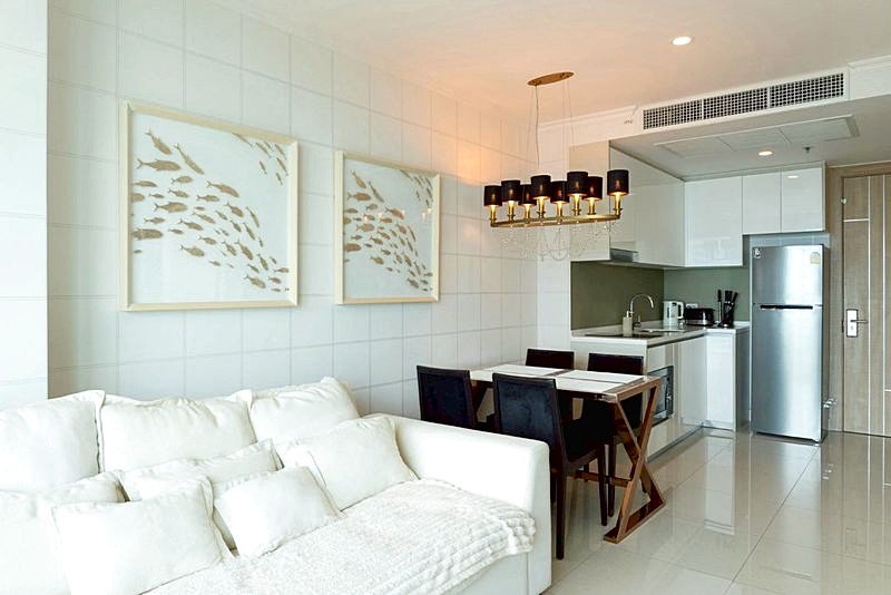 Open-plan living area with a white sofa, dining table, and a compact kitchen; a chandelier hangs over the table.