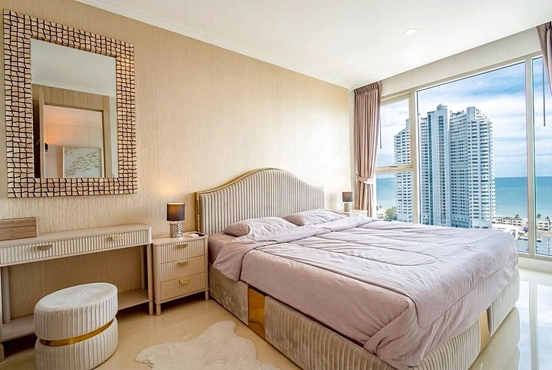 Hotel bedroom with a large bed, cream headboard, and soft lavender bedding; nightstands with lamps, dresser with mirror, and a round ottoman. Floor-to-ceiling windows show ocean and high-rise buildings.