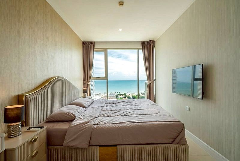 Hotel bedroom with a large window overlooking the ocean, a king-size bed with pink bedding, and a wall-mounted TV on the right.