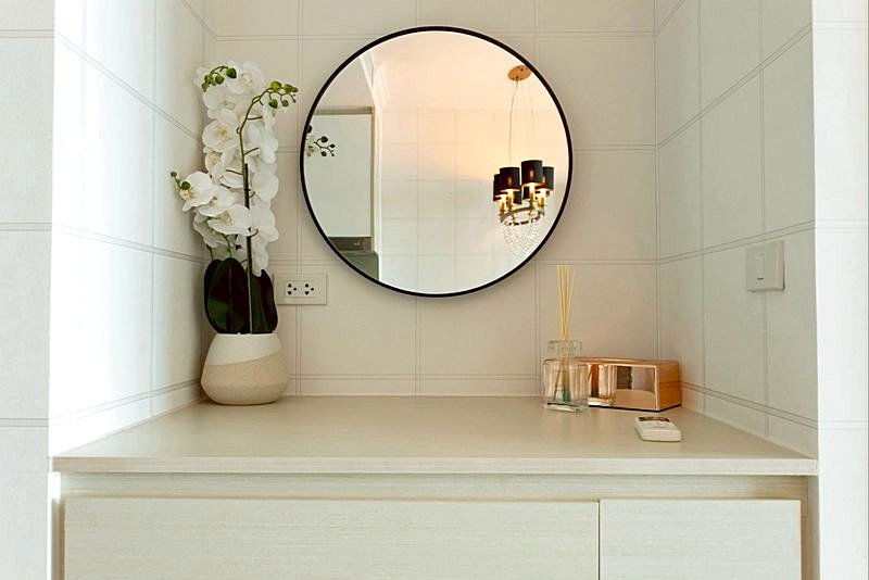 Round mirror above a white bathroom vanity, with a vase of white orchids on the left and a reed diffuser on the right.