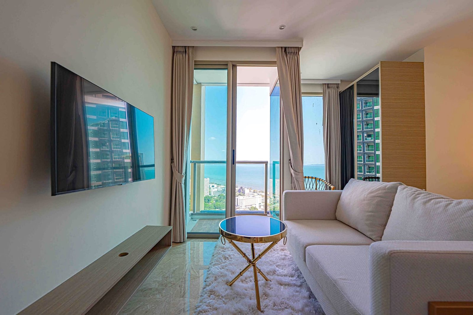 Riviera_Ocean_Drive_Studio_Rent_Rdl360 Living room with a wall-mounted TV on the left, beige curtains, and a glass-top round coffee table in front of a white sofa; a city view through large glass doors leads to a balcony.