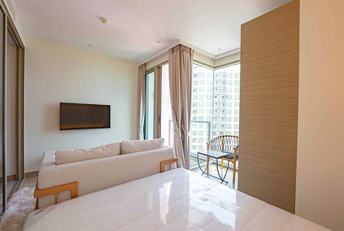 Bright living room with a wall-mounted TV, light sofa, and large windows overlooking city buildings; beige curtains frame a balcony.