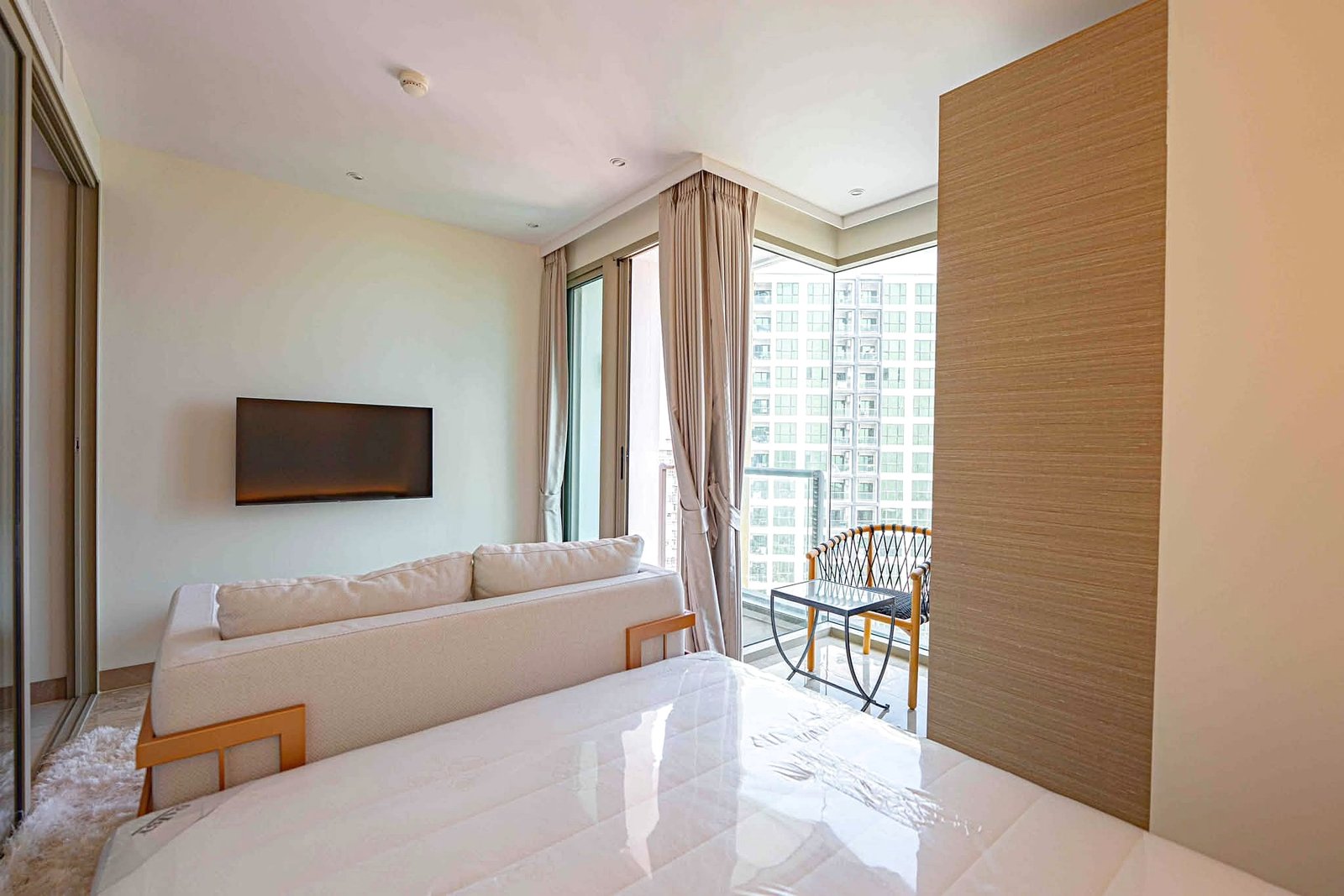 Riviera_Ocean_Drive_Studio_Rent_Rdl360_2 Bright living room with a wall-mounted TV, light sofa, and large windows overlooking city buildings; beige curtains frame a balcony.