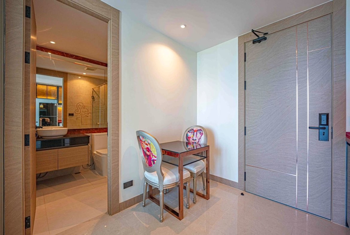 Interior of a modern hotel suite: left doorway to a bathroom with a vanity and mirror, center a small wooden table with two colorful upholstered chairs, and a tall paneled wardrobe on the right in neutral tones.