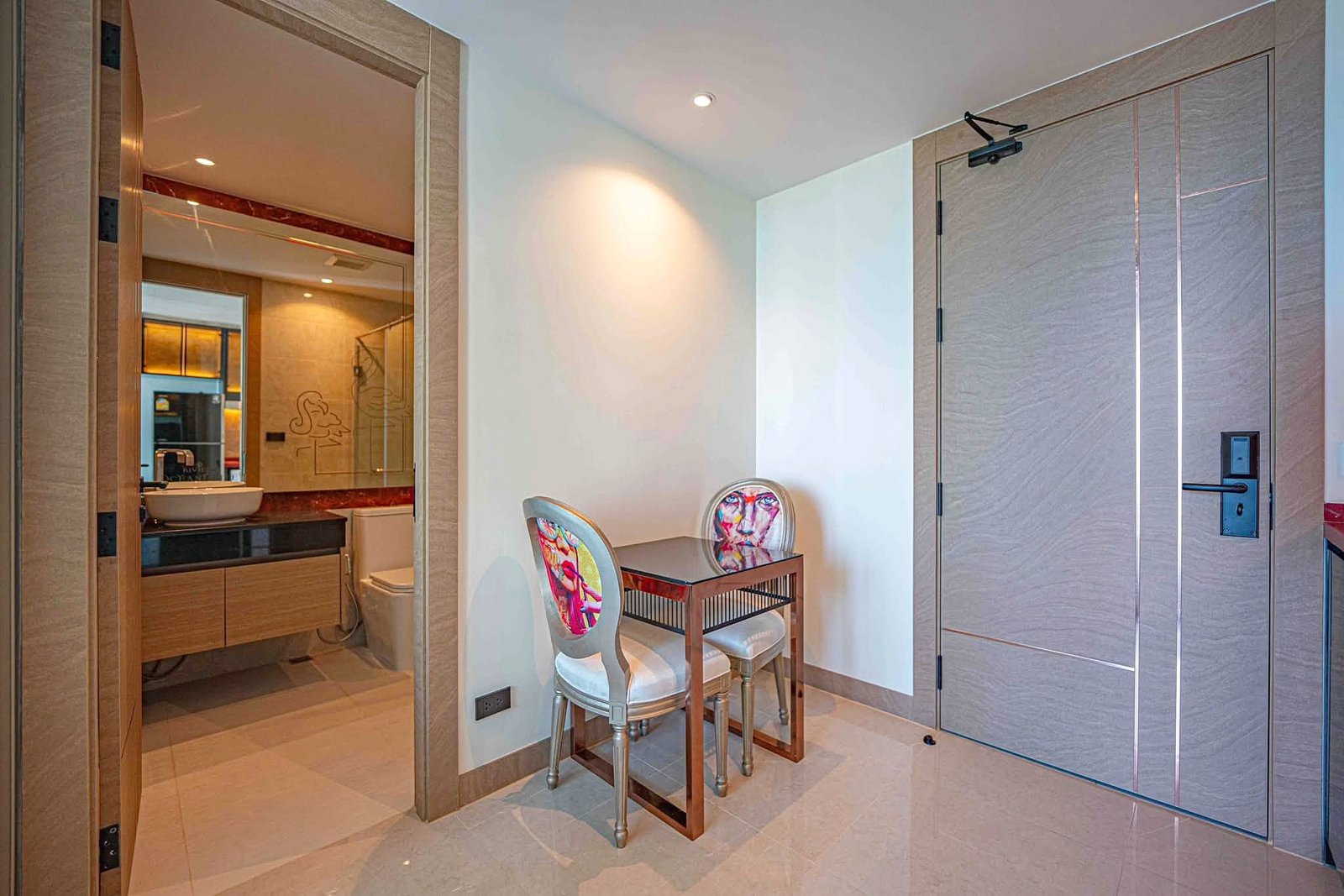 Riviera_Ocean_Drive_Studio_Rent_Rdl360_9 Interior of a modern hotel suite: left doorway to a bathroom with a vanity and mirror, center a small wooden table with two colorful upholstered chairs, and a tall paneled wardrobe on the right in neutral tones.