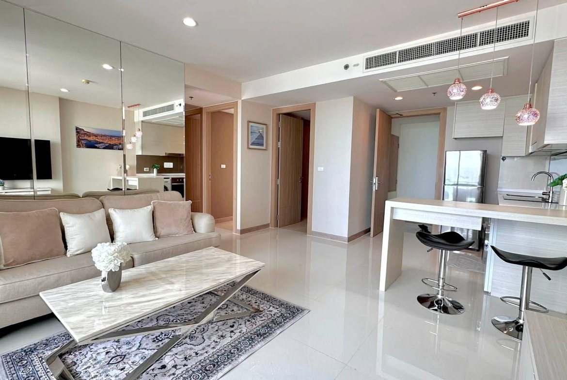 Open-concept living area with beige sofa, marble coffee table, mirrored wall, and a bright kitchen with a bar-height counter and stools.