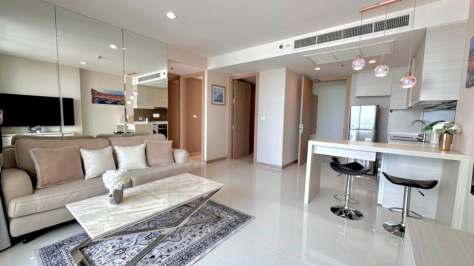 Riviera_Wongamat_2bed_Dlc640 Open-concept living area with beige sofa, marble coffee table, mirrored wall, and a bright kitchen with a bar-height counter and stools.