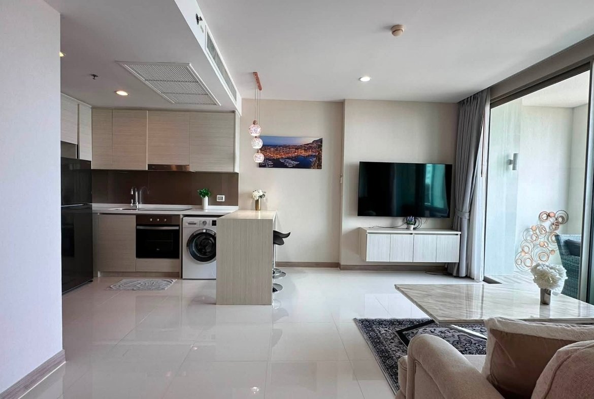 Open-plan living area with a modern kitchen on the left, island, washing machine, and a wall-mounted TV opposite the glass balcony doors.