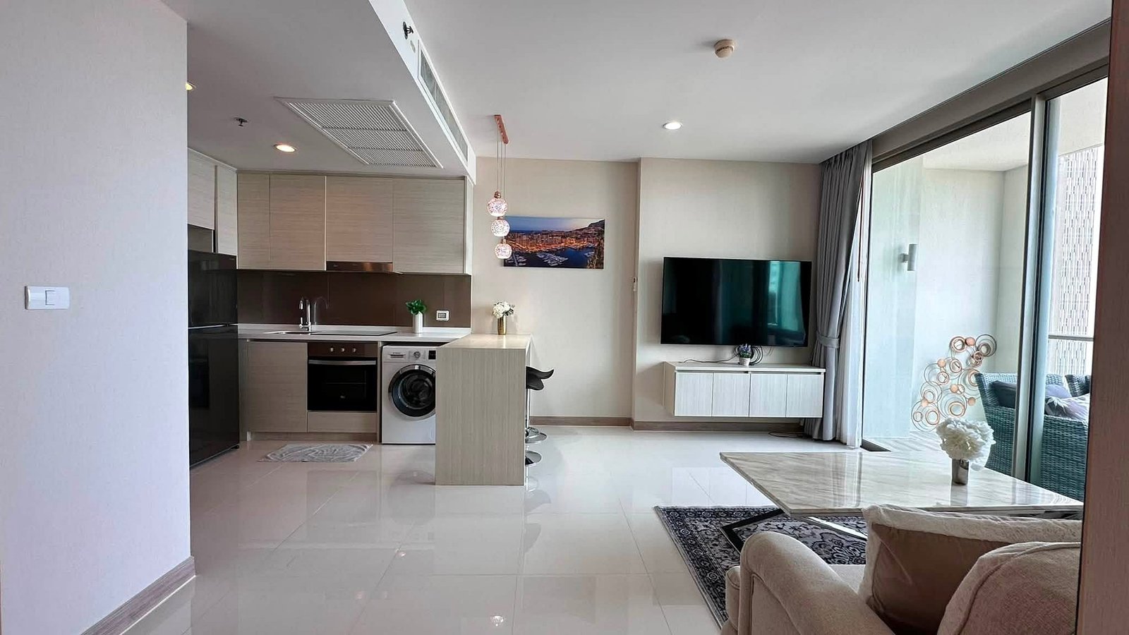 Riviera_Wongamat_2bed_Dlc640_10 Open-plan living area with a modern kitchen on the left, island, washing machine, and a wall-mounted TV opposite the glass balcony doors.