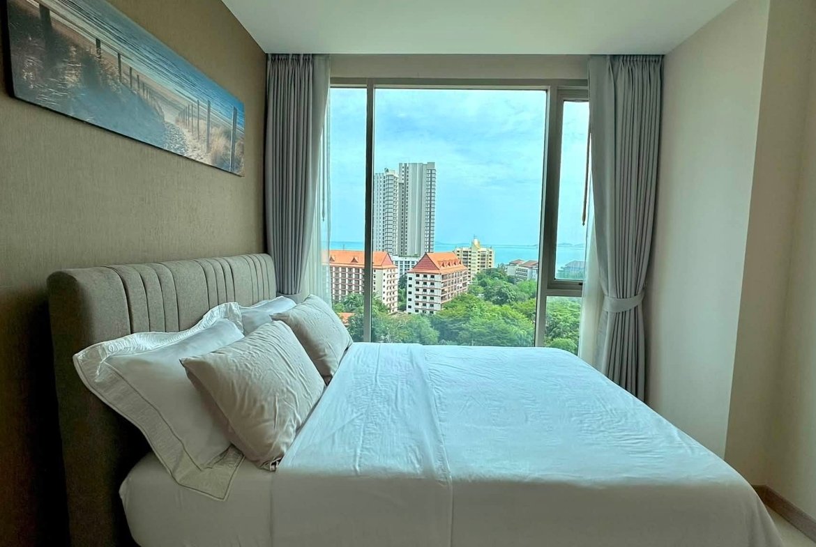 Hotel bedroom with a white bed and pillows, and a large window revealing city-highrises and blue sea beyond.