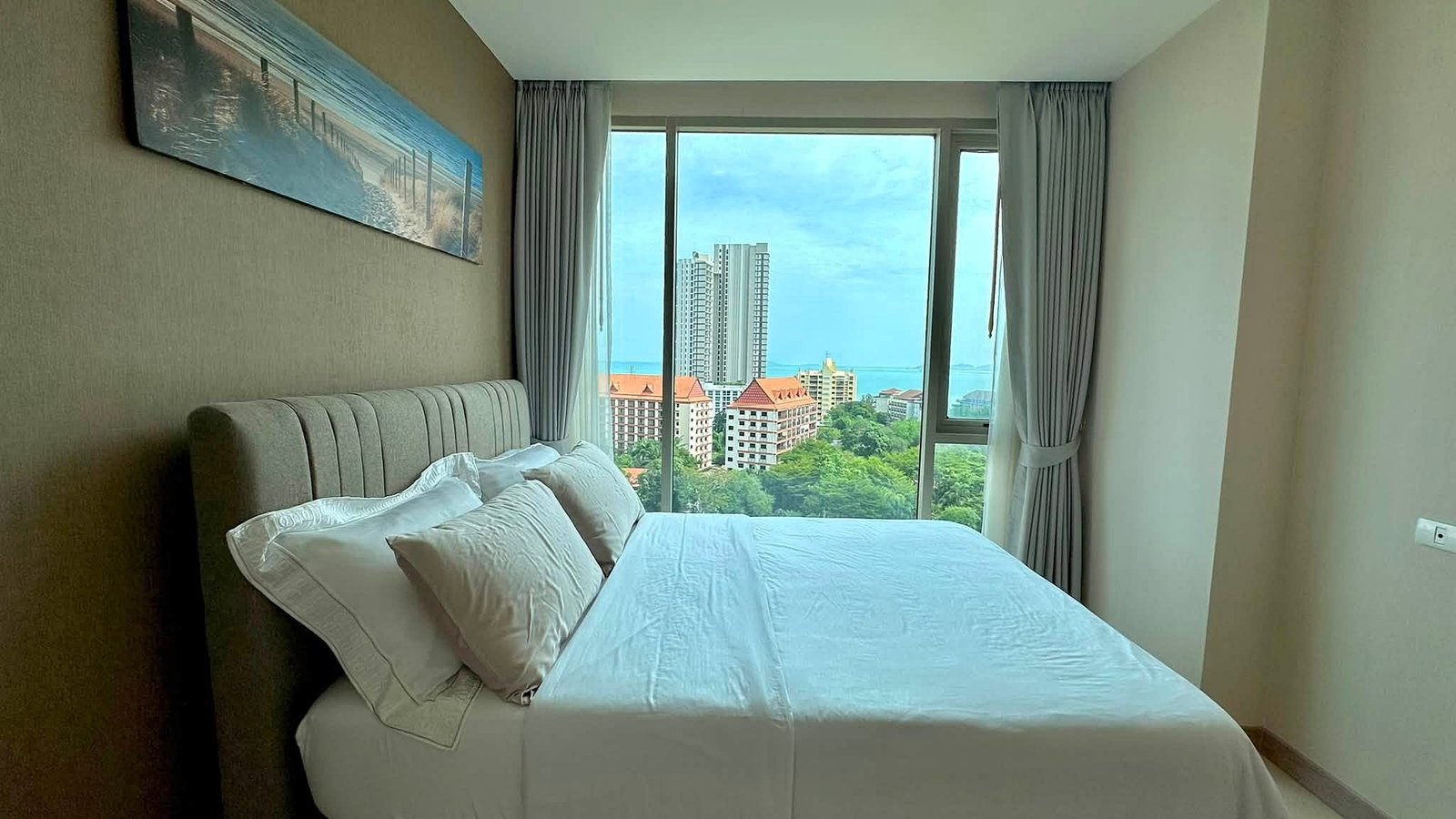 Riviera_Wongamat_2bed_Dlc640_12 Hotel bedroom with a white bed and pillows, and a large window revealing city-highrises and blue sea beyond.