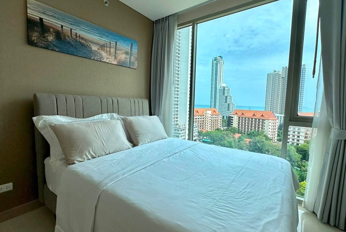 Hotel bedroom with white bedding and a gray upholstered headboard, large windows reveal city skyscrapers and the ocean beyond.