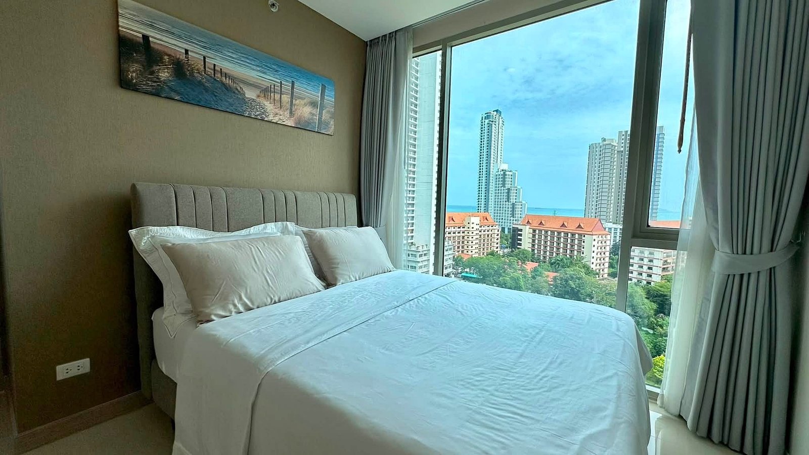 Riviera_Wongamat_2bed_Dlc640_13 Hotel bedroom with white bedding and a gray upholstered headboard, large windows reveal city skyscrapers and the ocean beyond.