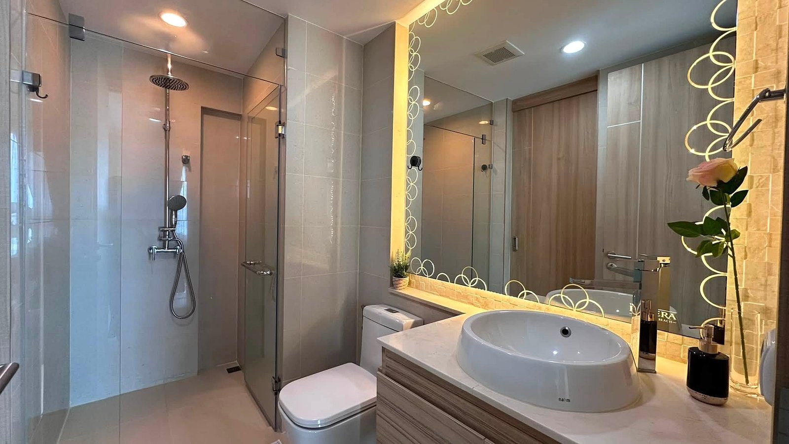 Riviera_Wongamat_2bed_Dlc640_15 Modern bathroom with glass shower enclosure, large circular-pattern mirror, and a round vessel sink on a wood vanity.