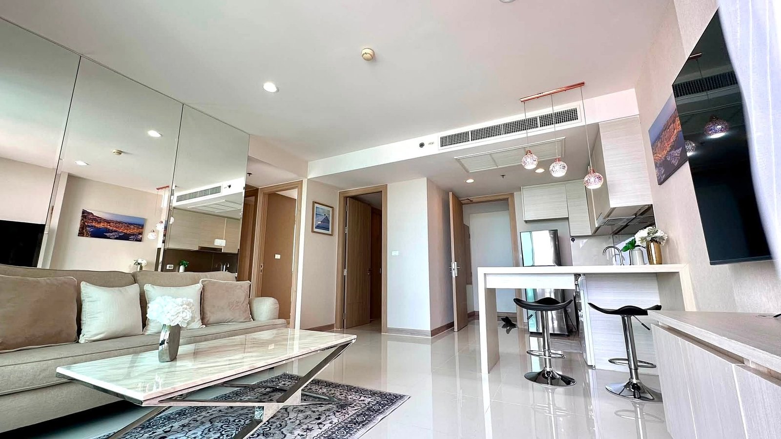 Riviera_Wongamat_2bed_Dlc640_18 Open-plan living area with a beige sofa, marble coffee table, mirrored wall, white kitchen island, and pendant lights.
