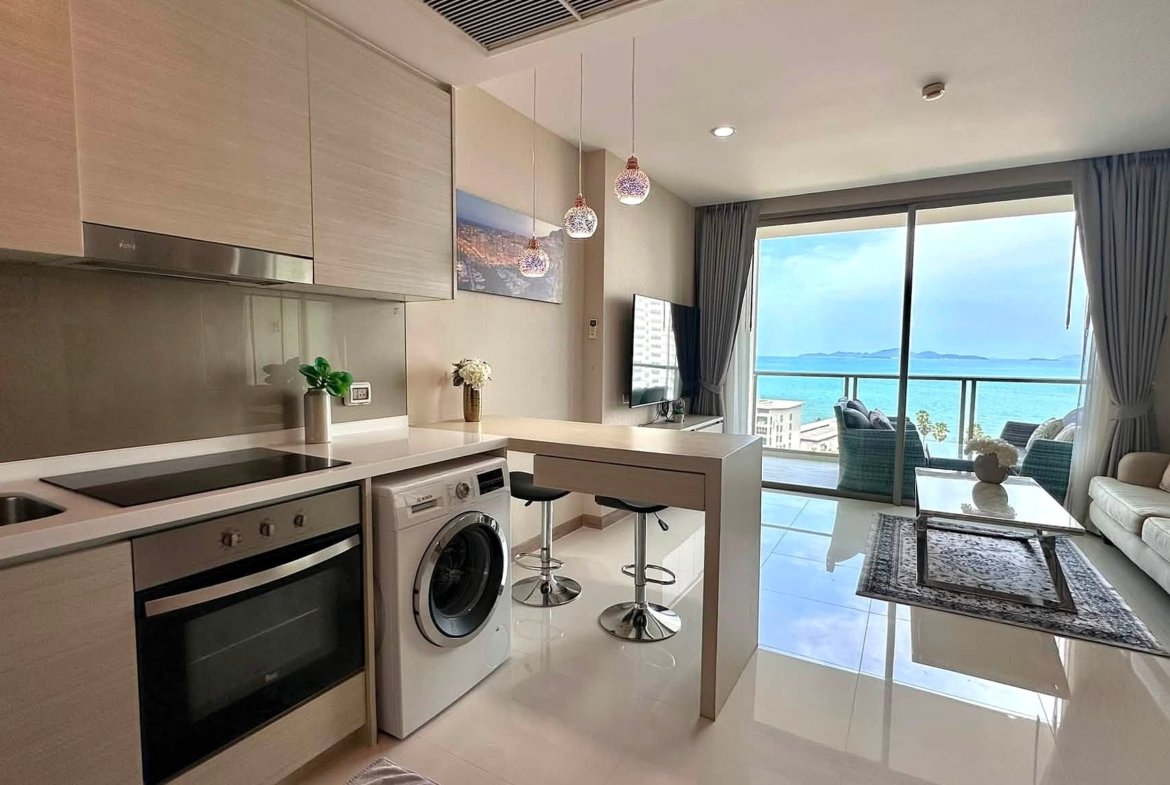 Open-plan kitchen with white countertops, built-in oven and washing machine, island bar, and a sea-view living area beyond.