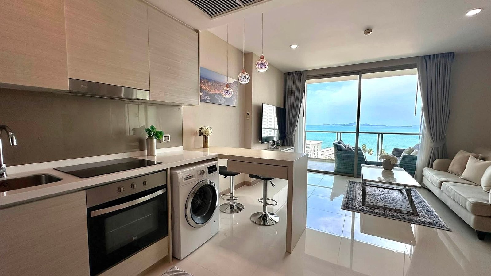 Riviera_Wongamat_2bed_Dlc640_2 Open-plan kitchen with white countertops, built-in oven and washing machine, island bar, and a sea-view living area beyond.