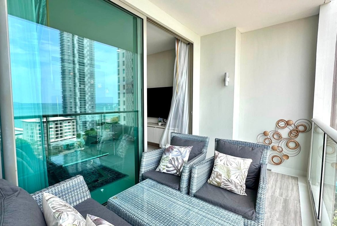 Balcony seating area with gray cushions and tropical-pattern pillows, overlooking high-rise buildings and the ocean.