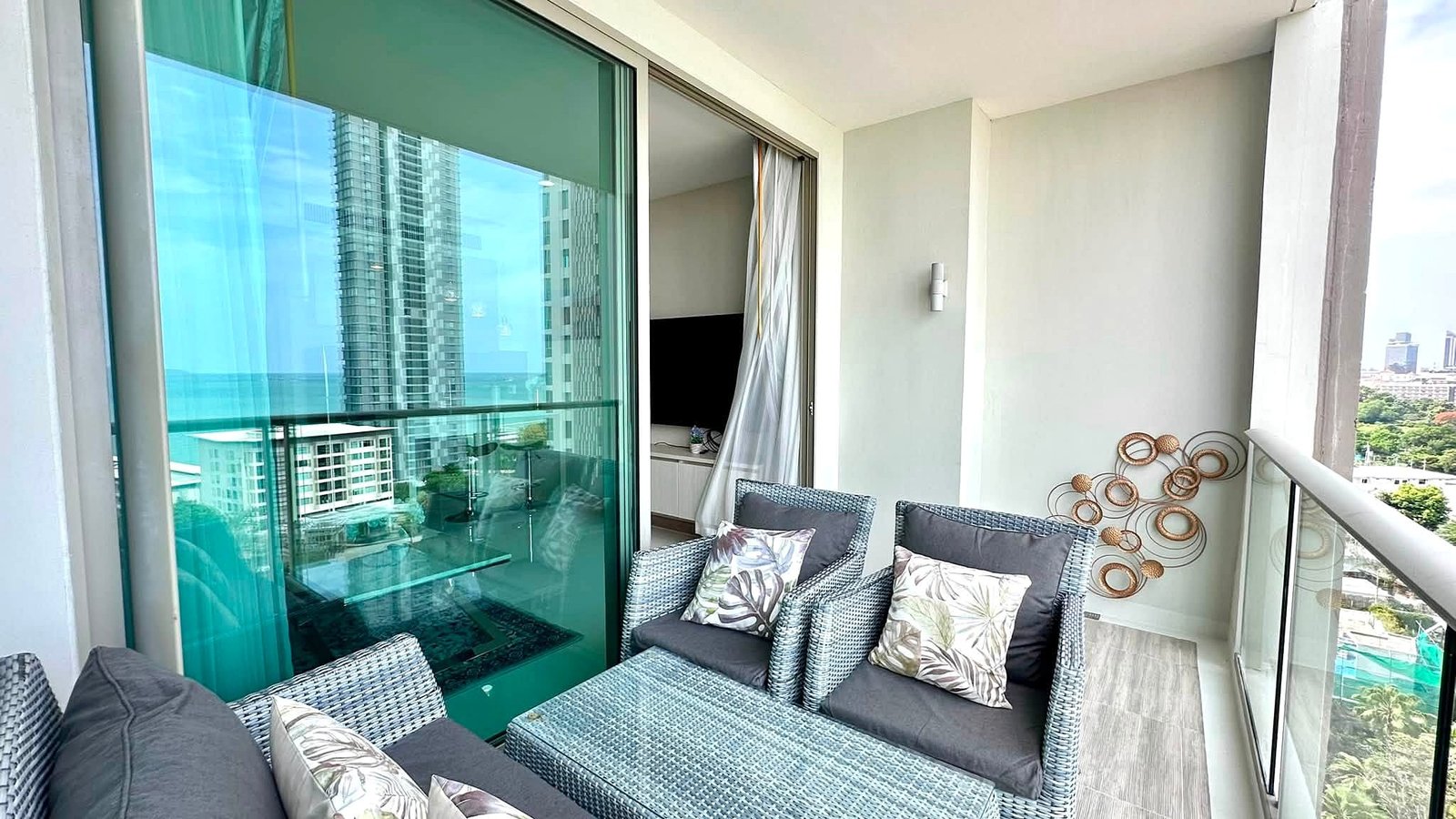 Riviera_Wongamat_2bed_Dlc640_21 Balcony seating area with gray cushions and tropical-pattern pillows, overlooking high-rise buildings and the ocean.