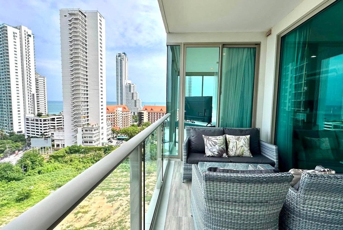 Balcony seating with wicker sofa and glass table overlooking modern high-rise buildings and the ocean.