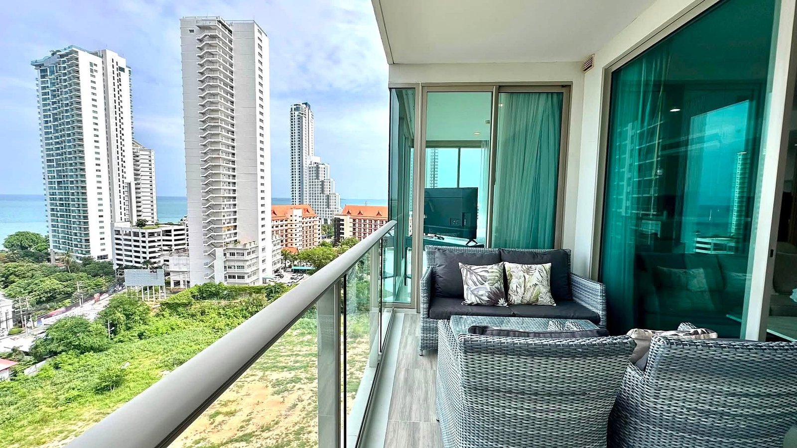 Riviera_Wongamat_2bed_Dlc640_22 Balcony seating with wicker sofa and glass table overlooking modern high-rise buildings and the ocean."
