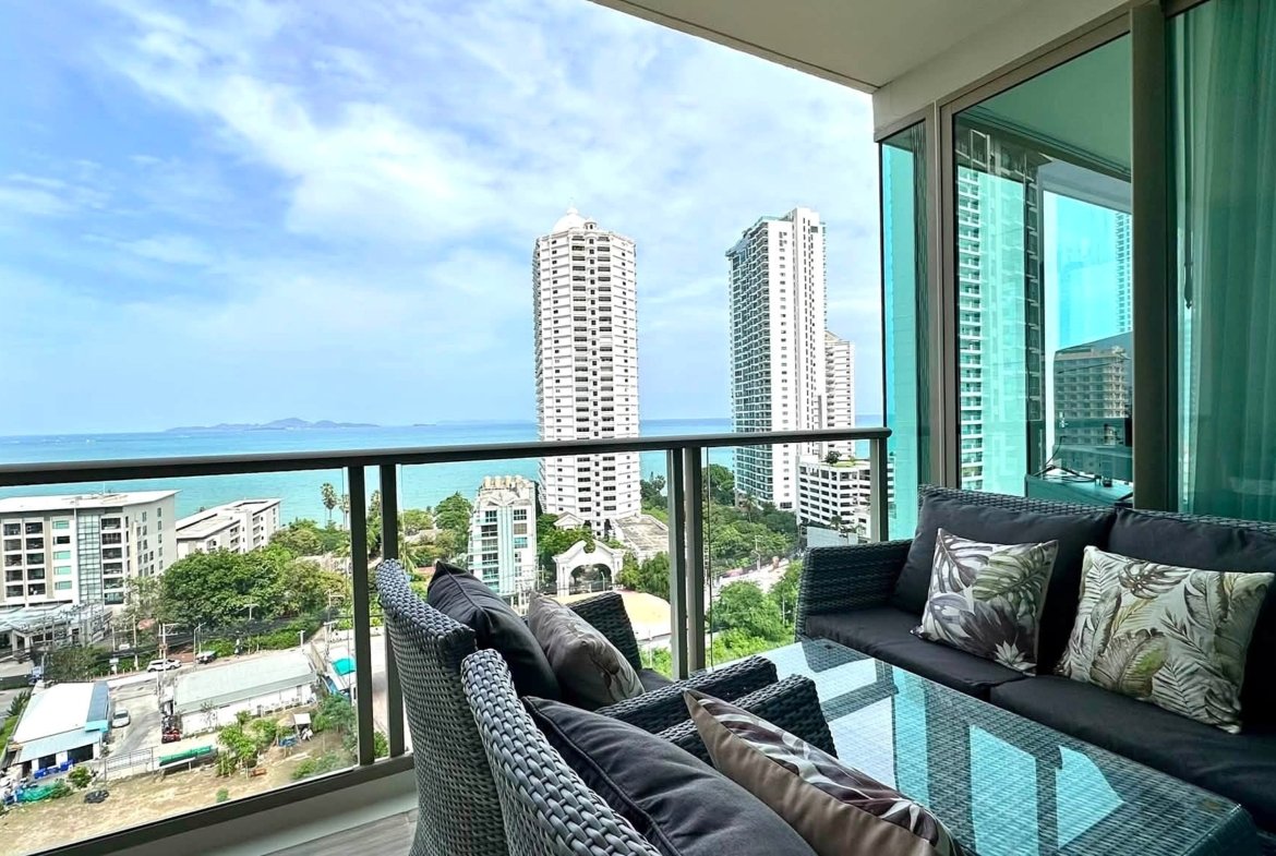 Balcony seating area with dark wicker couches and tropical cushions, overlooking white high-rise towers and the ocean.