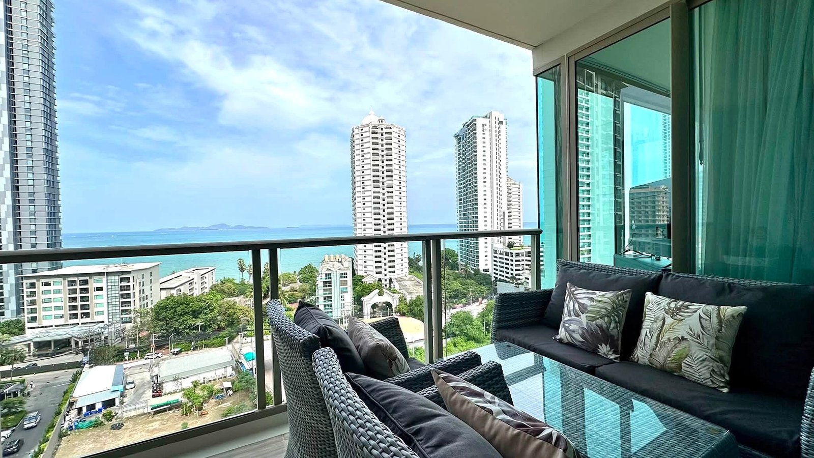 Riviera_Wongamat_2bed_Dlc640_23 Balcony seating area with dark wicker couches and tropical cushions, overlooking white high-rise towers and the ocean.
