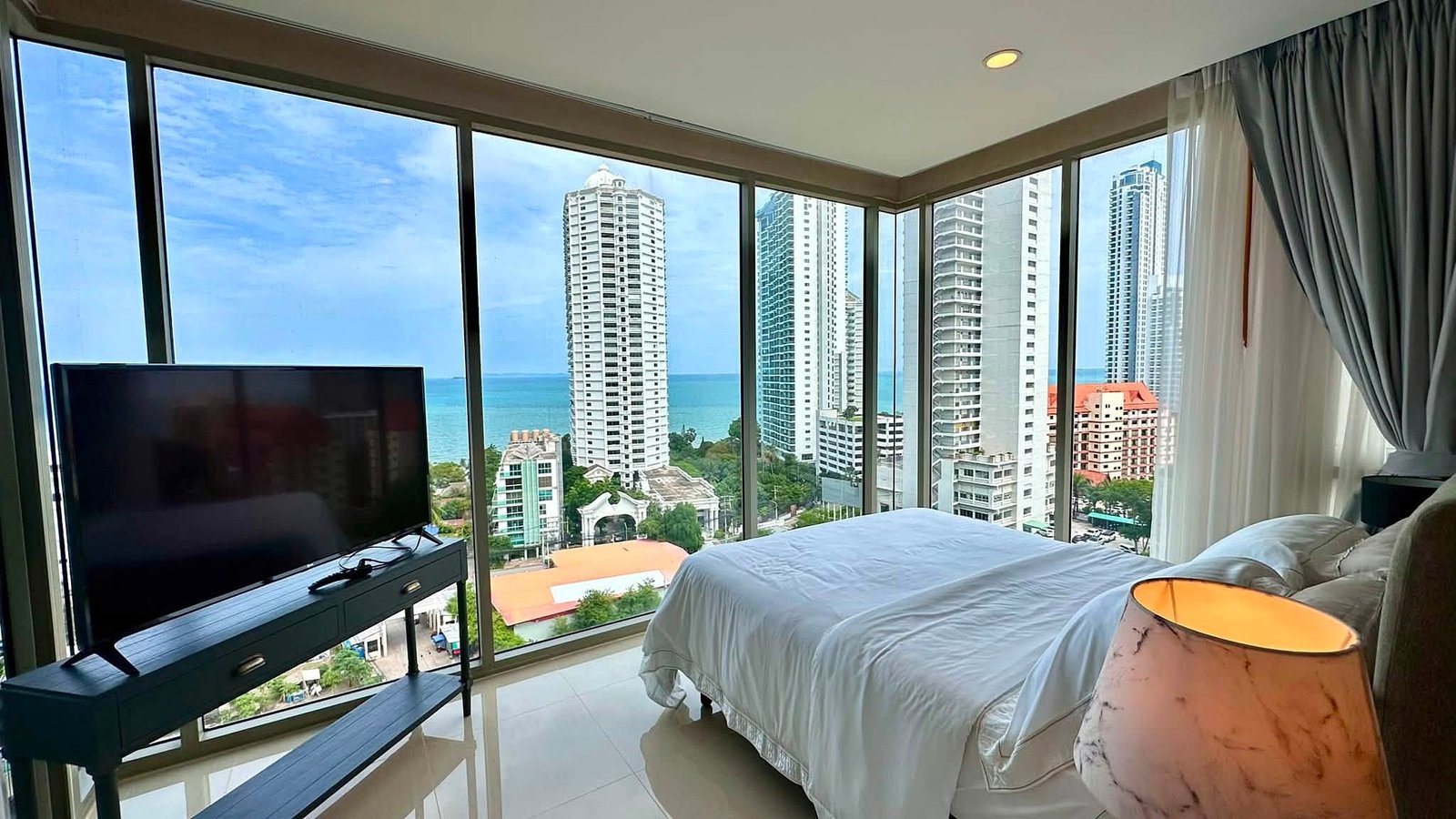 Riviera_Wongamat_2bed_Dlc640_3 Hotel room with floor-to-ceiling windows overlooking the ocean and surrounding high-rise buildings at sea bluff.