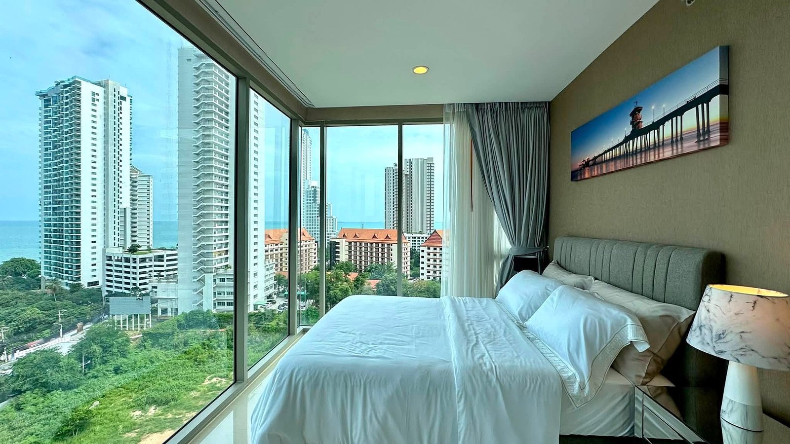 Riviera_Wongamat_2bed_Dlc640_4 Hotel room with floor-to-ceiling windows showing city high-rises and the ocean beyond.
