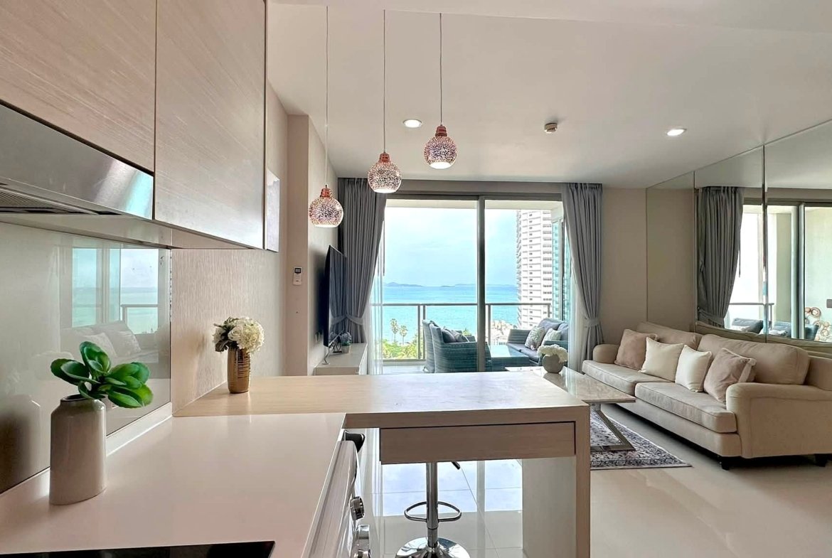 Bright open-plan kitchen and living room with white glossy surfaces, beige sofa, and a sea-view balcony beyond glass doors.