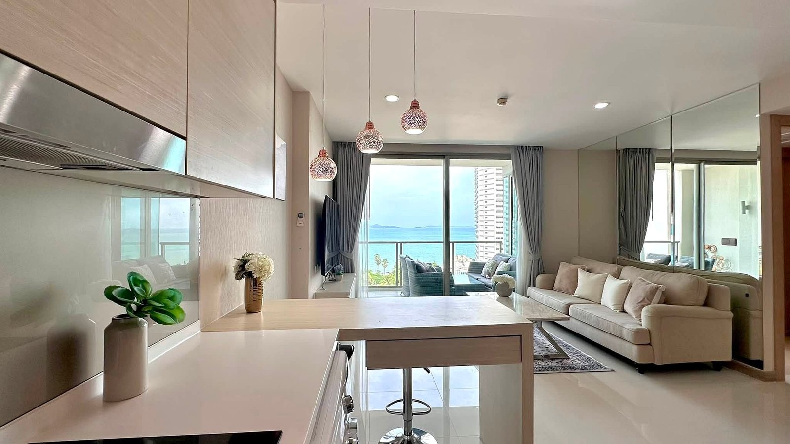Riviera_Wongamat_2bed_Dlc640_9 Bright open-plan kitchen and living room with white glossy surfaces, beige sofa, and a sea-view balcony beyond glass doors.