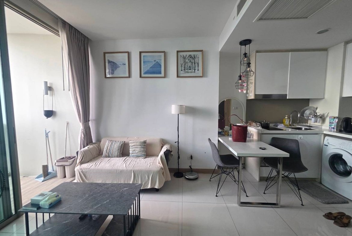 Open-plan living space with a beige sofa, three framed pictures on the wall, and a white kitchen with a dining table nearby.