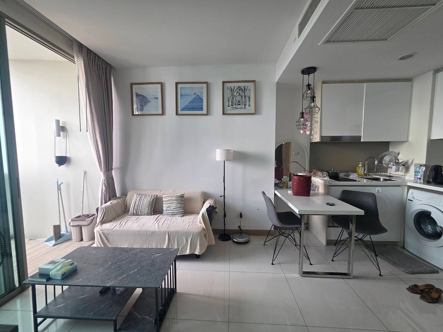 Riviera_Wongamat_B1209 Open-plan living space with a beige sofa, three framed pictures on the wall, and a white kitchen with a dining table nearby.